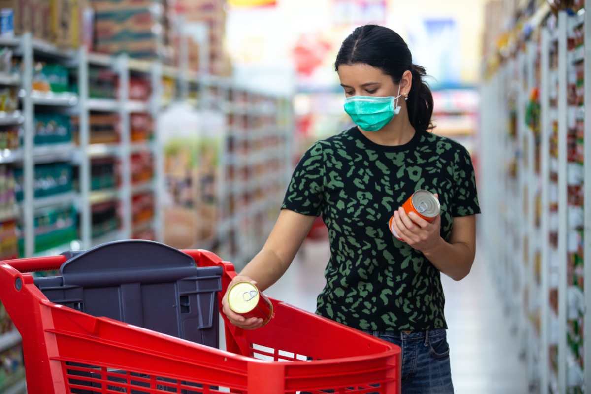 female wears medical mask against coronavirus while grocery shopping in supermarket or store- health, safety and pandemic concept - young woman wearing protective mask and stockpiling food
