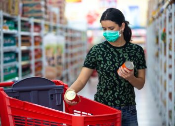 female wears medical mask against coronavirus while grocery shopping in supermarket or store- health, safety and pandemic concept - young woman wearing protective mask and stockpiling food