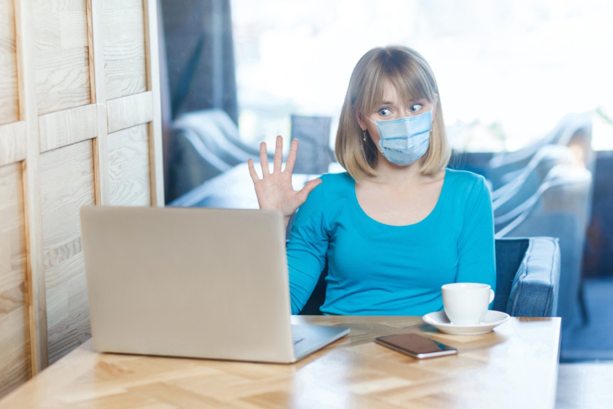 woman with surgical medical mask is sitting and working on laptop and greeting on video call conference