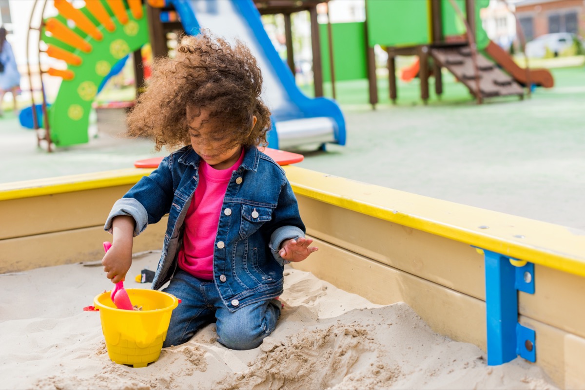 curly african american little child playing with plastic scoop and bucket in sandbox at playground