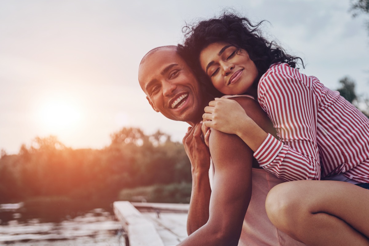 Happy young couple embracing and smiling while sitting on the pier near the lake