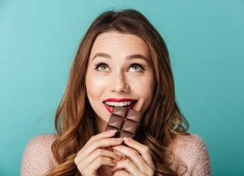 Portrait of a delighted brown haired woman with bright makeup eating chocolate bar