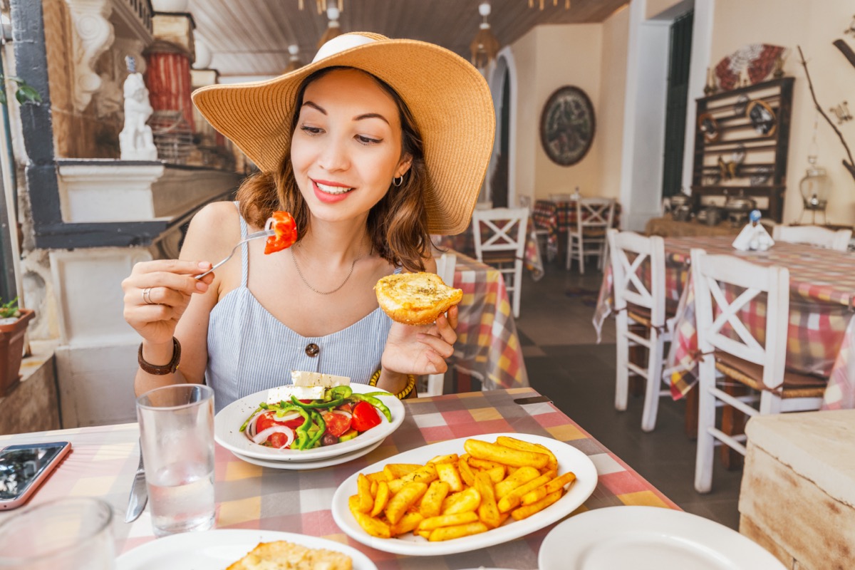 Happy asian woman in big hat having meal with greek salad Horiatiki in restaurant. Greece cuisine concept