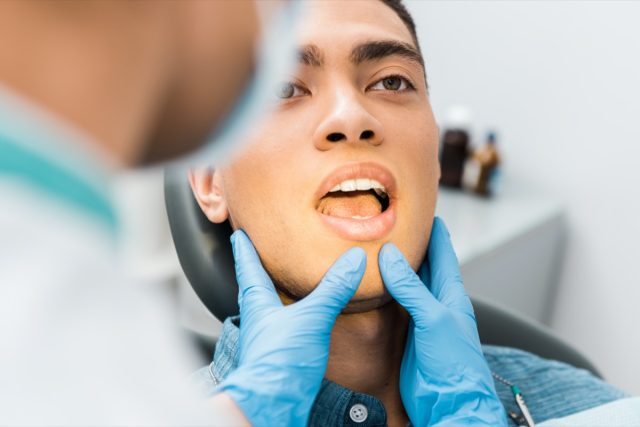 Female dentist hands in latex gloves touching face of a man with open mouth.