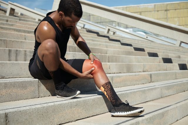 runner with athletic body wearing black running shoes, sitting on steps on concrete stair, clutching injured knee in excruciating pain