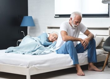 Worried senior depressed man sitting in bed and suffering from insomnia while his wife sleep