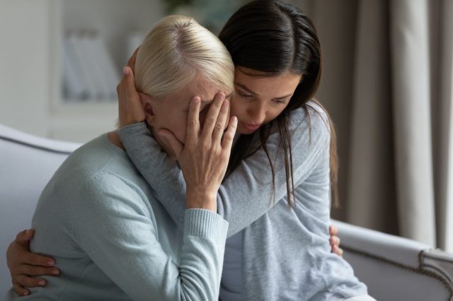 Empathic young lady embracing soothing crying depressed elder mommy, sitting together at home