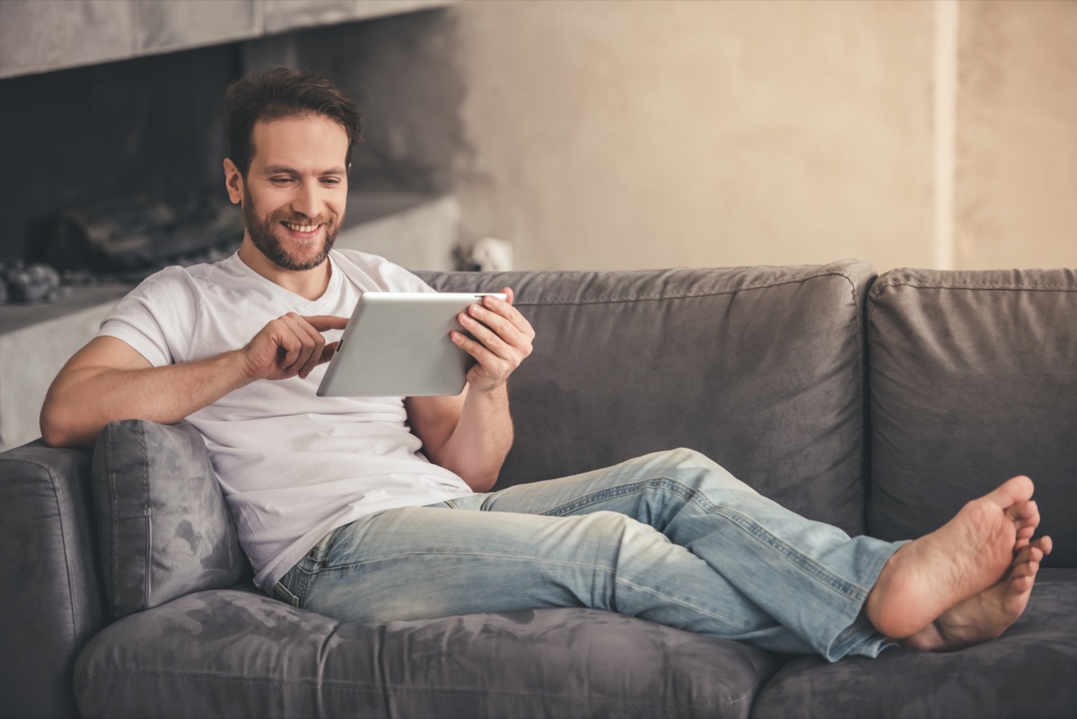 Handsome man is using a digital tablet and smiling while resting on couch at home