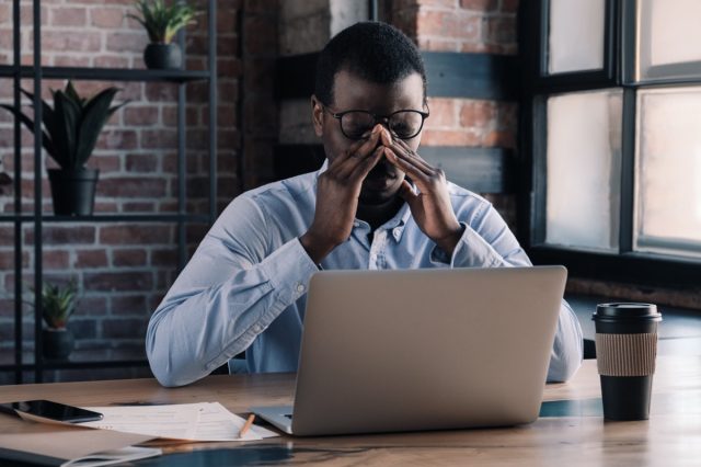 Tired african business man massaging his nose with eyes closed, sitting at his desk in office