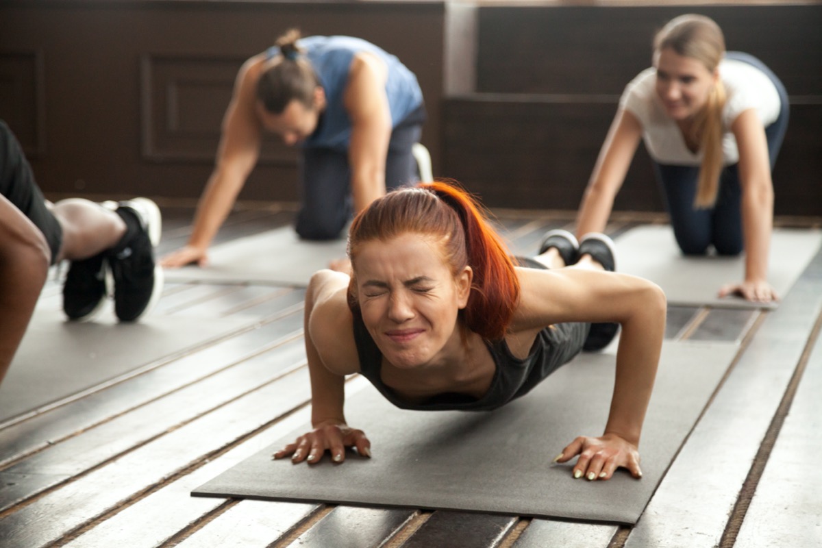 woman with painful face expression doing hard difficult plank fitness exercise or push press ups feeling pain in muscles at diverse group training class in gym