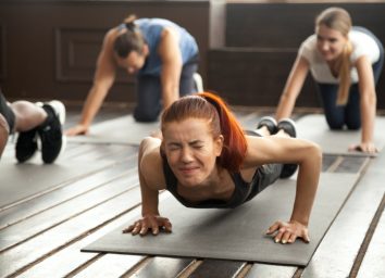 woman with painful face expression doing hard difficult plank fitness exercise or push press ups feeling pain in muscles at diverse group training class in gym
