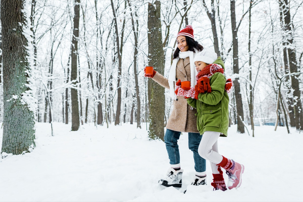 mother and cute daughter walking and holding red cups between trees and looking at snow in snowy forest