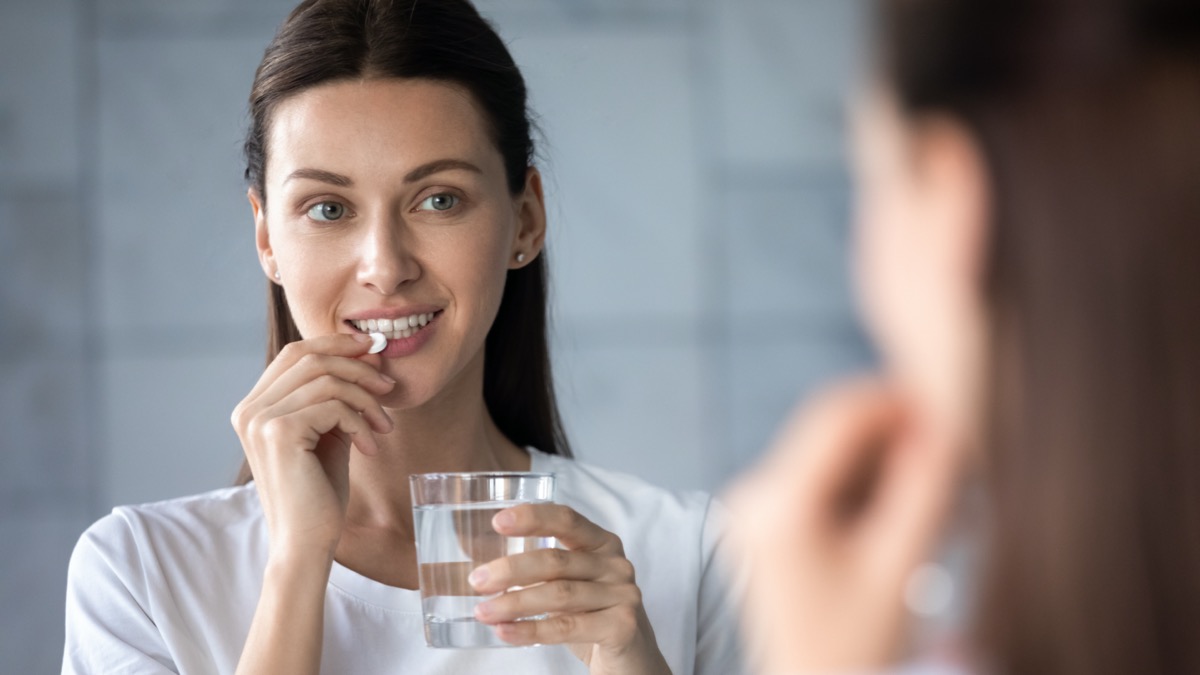 Young healthy woman holding pill glass of water looking in mirror