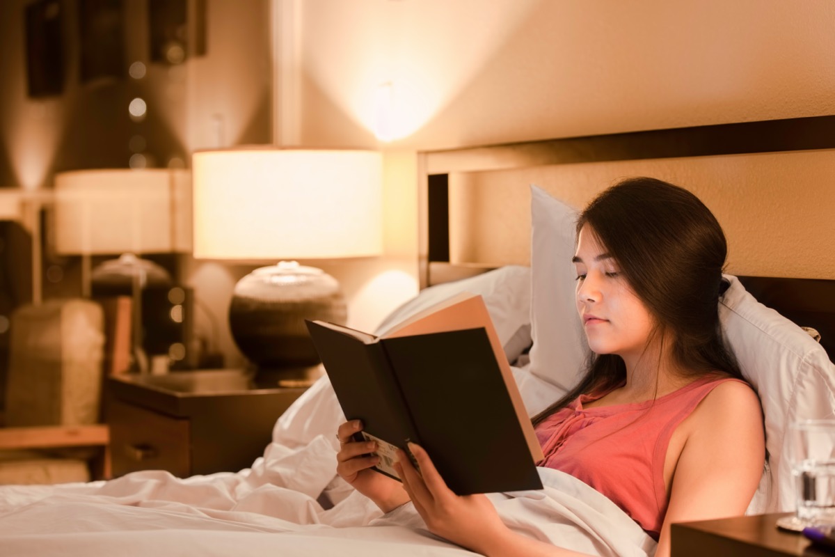 Asian Caucasian teen girl reading book at night in bed with yellow lamp light on walls