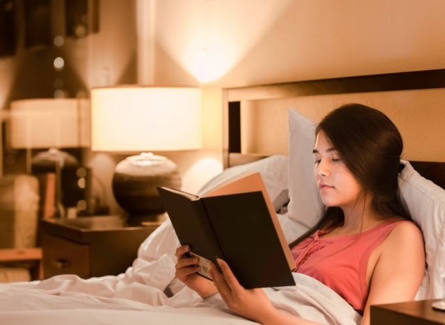 Asian Caucasian teen girl reading book in bed at night with yellow lamp light on walls