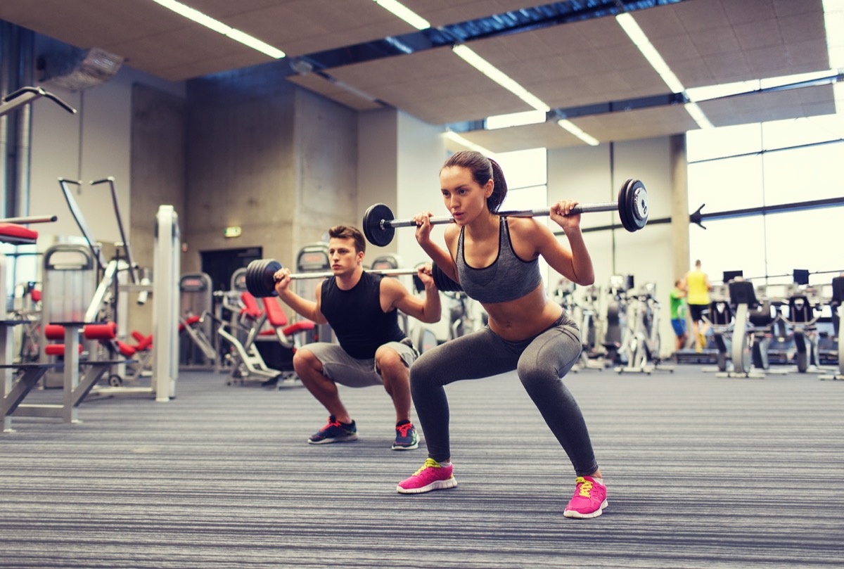 young man and woman with barbell flexing muscles and making shoulder press squat in gym