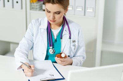 physician medicine doctor or pharmacist sitting at worktable, holding jar of pills in hands and writing prescription on special form.