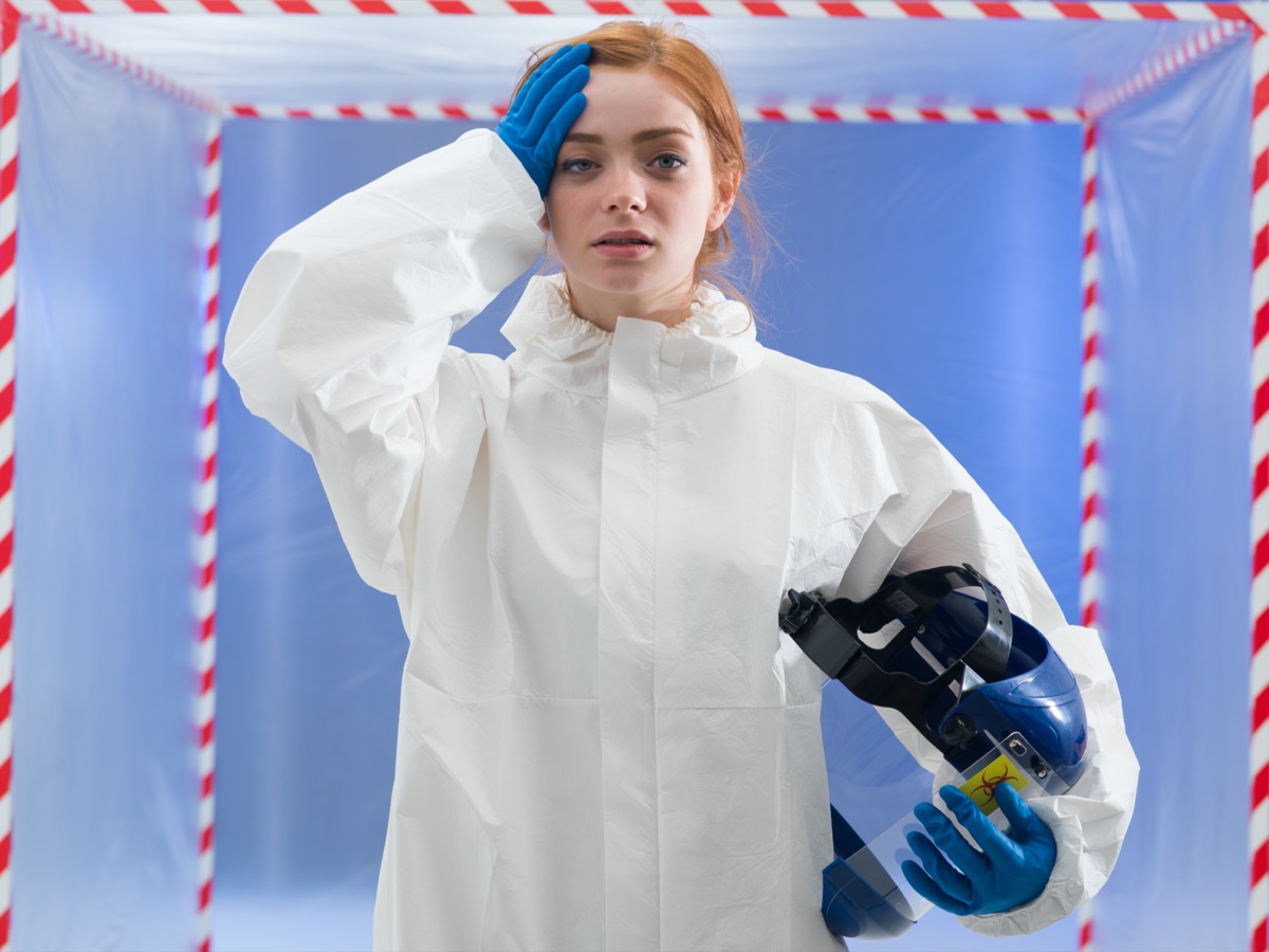 Exhausted female biohazard scientist dressed in a biohazard suit holding her helmet mask in one hand and her forehead with the other
