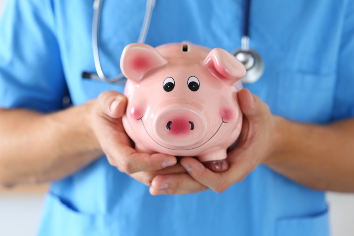 Male medicine doctor wearing blue uniform holding happy funny smiling piggybank in hands closeup. Medical service economy, health care savings and insurance concept