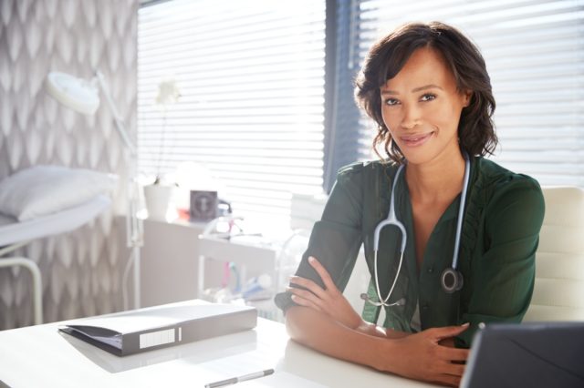 Portrait Of Smiling Female Doctor With Stethoscope Sitting Behind Desk In Office