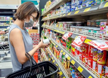 woman wearing protective face mask holding shopping basket and choosing food can product