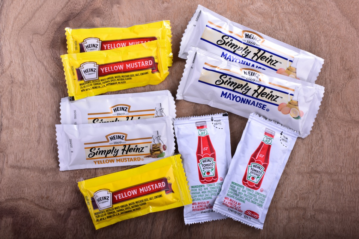 Various single serve packets of Heinz products including ketchup mayo and yellow mustard on a cherry wood countertop.