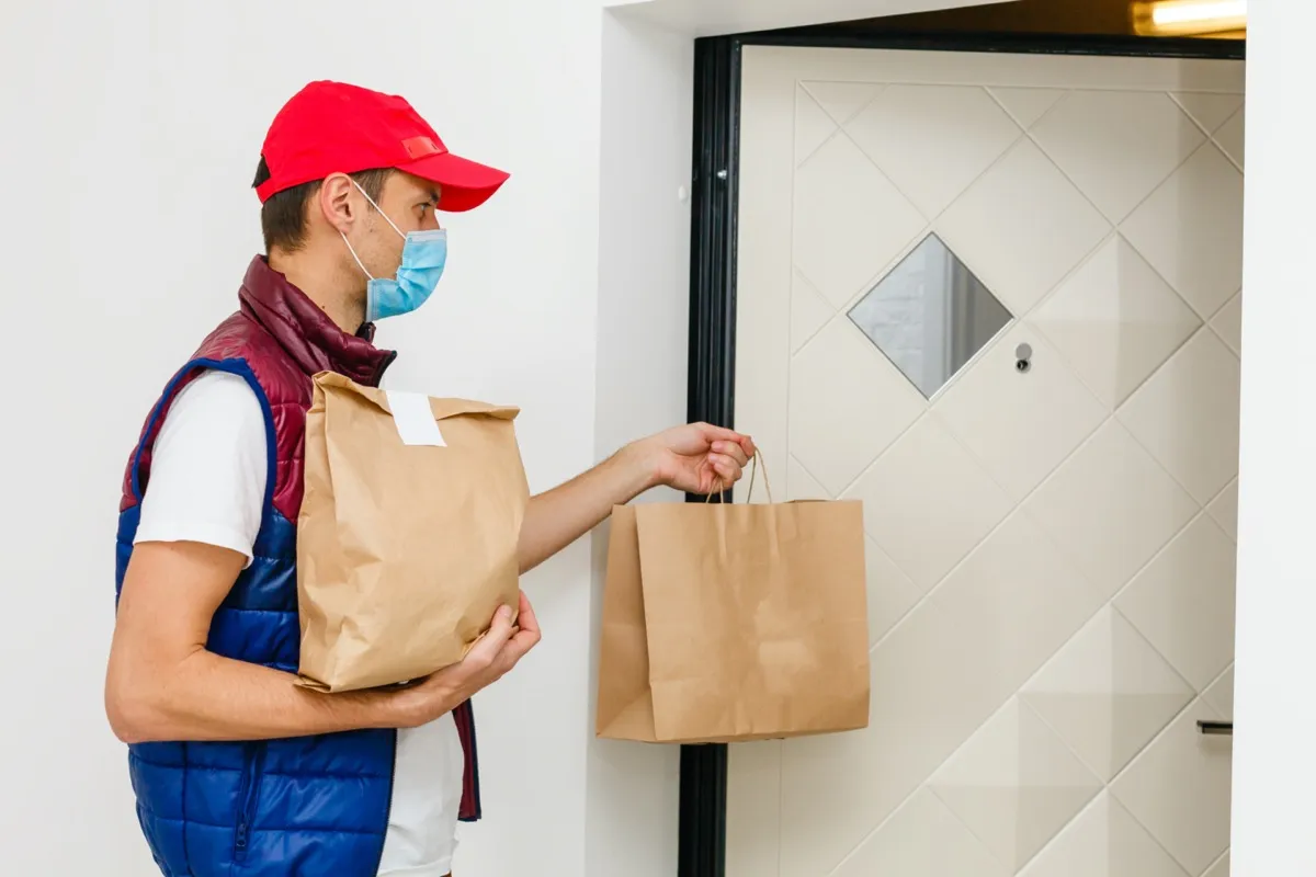 Delivery man holding paper bag with food on white background, food delivery man in protective mask