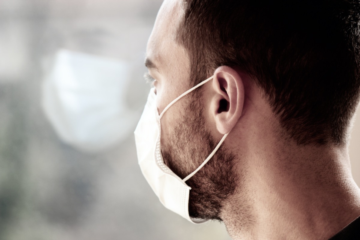 Photo of a young white man with medical face mask looking out of the window during coronavirus quarantine. Reflection of his face in the window.