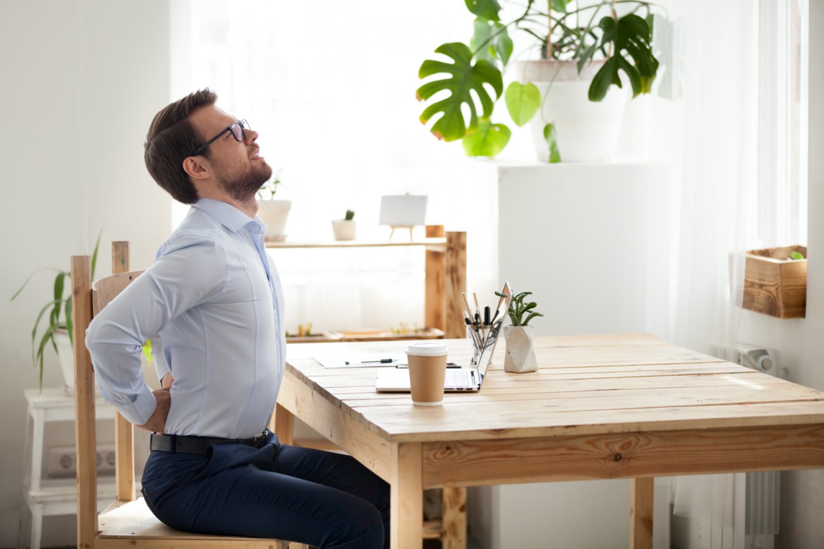 Tired millennial office worker stretch in chair suffer from sitting long in incorrect posture, male employee have back pain or spinal spasm working in uncomfortable position