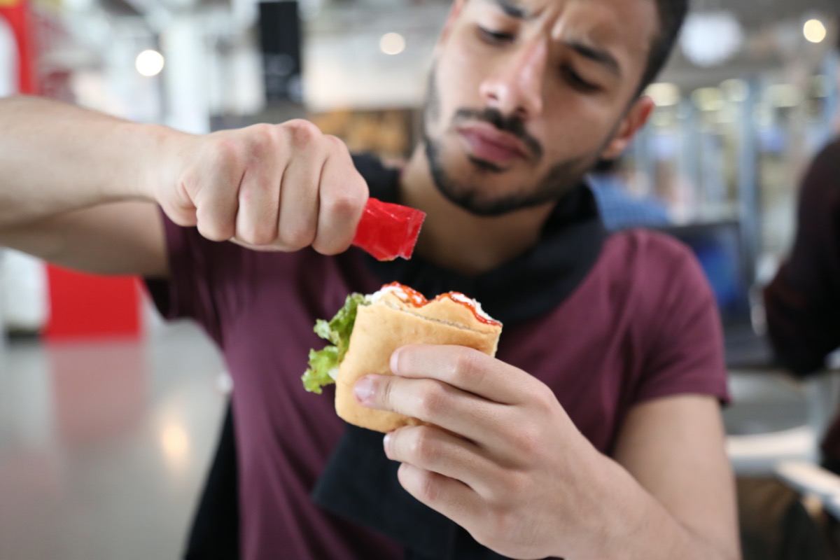 Hungry man sitting in a restaurant, holding a ketchup packet adding it to his sandwich