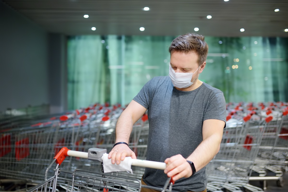 Man wearing disposable medical face mask wipes the shopping cart handle with a disinfecting cloth in supermarket