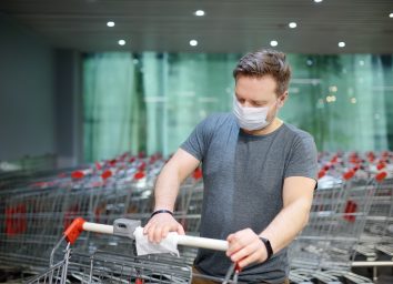 Man wearing disposable medical face mask wipes the shopping cart handle with a disinfecting cloth in grocery store