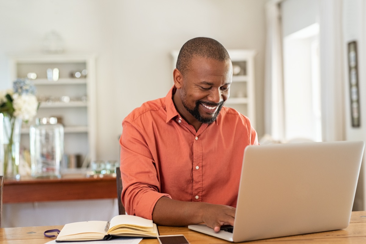 Smiling black man using laptop at home in living room. Happy mature businessman send email and working at home