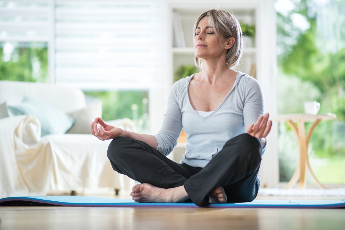 Middle aged woman sitting in the lotus position on a rug in her living room.  his eyes are closed.  she is in the foreground
