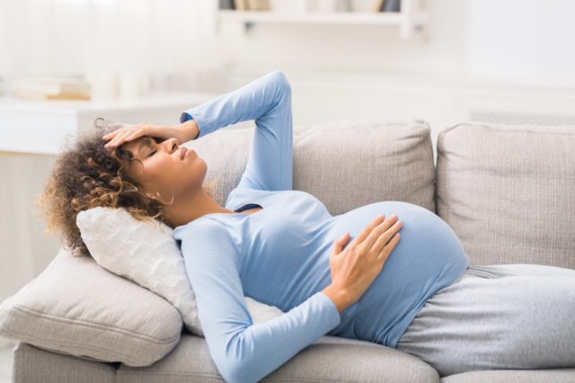 woman with headache lying on sofa, touching her belly and forehead