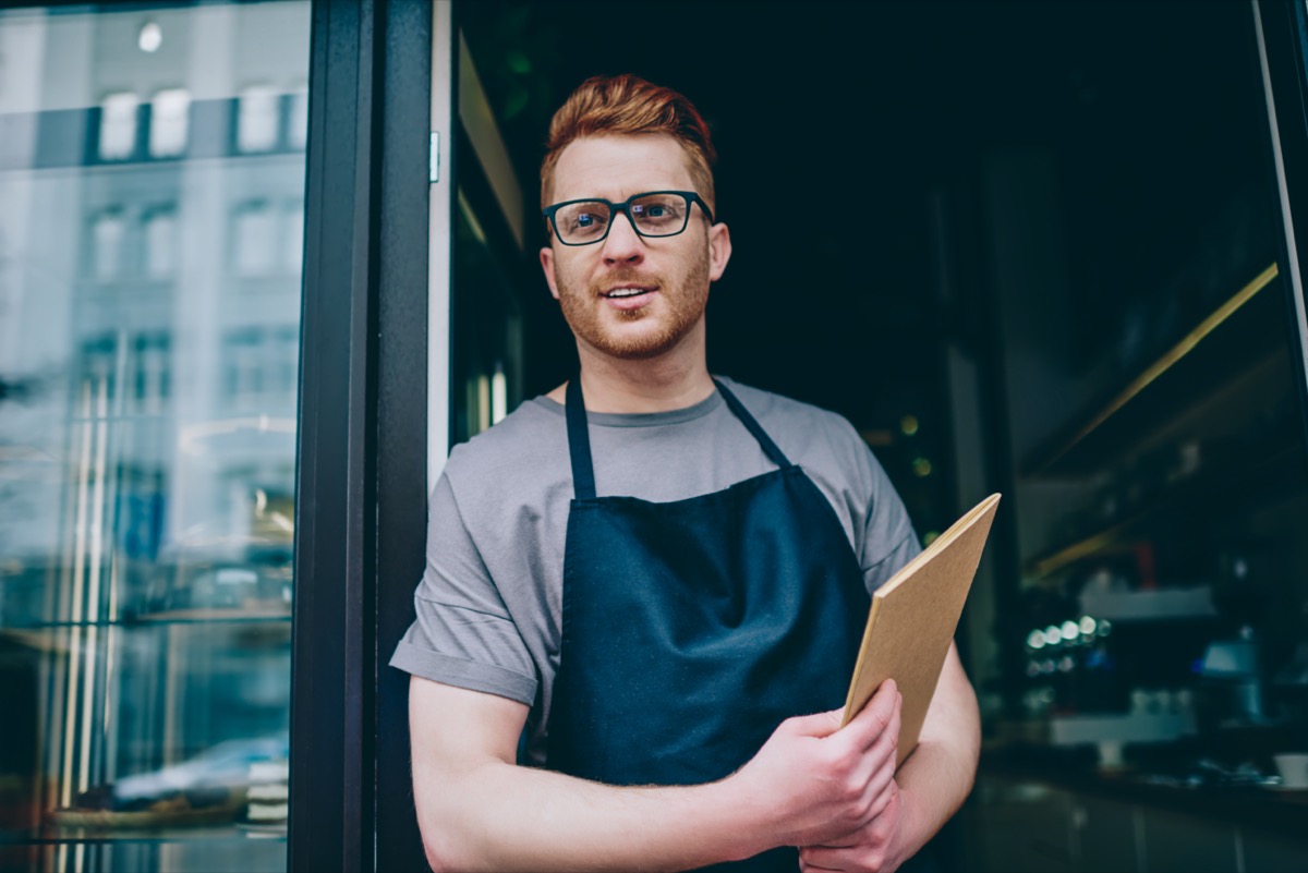 waiter in black apron holding menu and smiling while inviting guests to own bakery shop