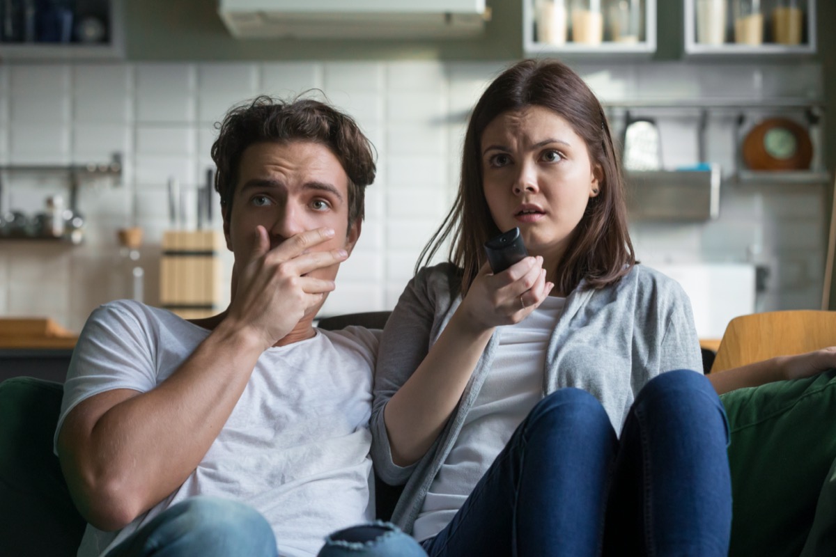 Scared millennial couple watching horror movie on tv holding remote control at home