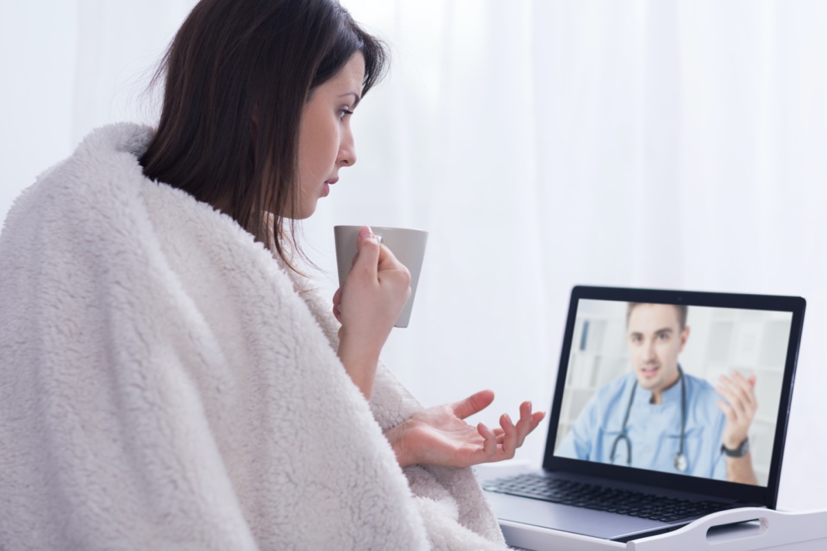 Shot of a woman covered with thick blanket, holding a white cup during on-line consultation with a GP
