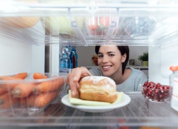 Smiling young woman having an unhealthy snack, she is taking a delicious pastry out of the fridge