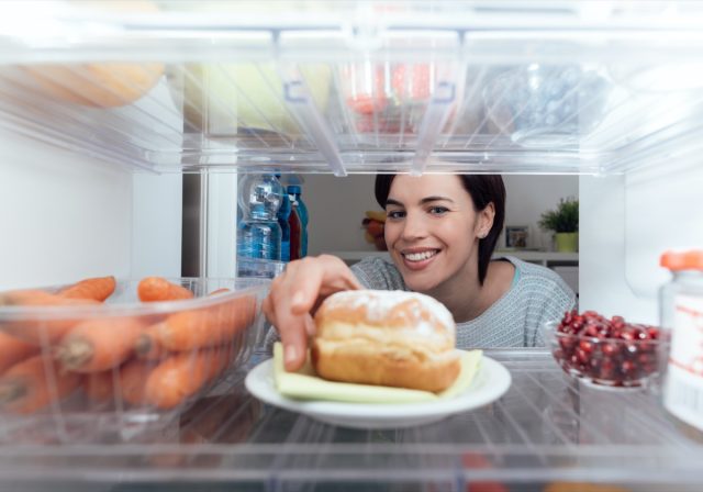 Smiling young woman eating an unhealthy snack, taking a delicious pie from the refrigerator