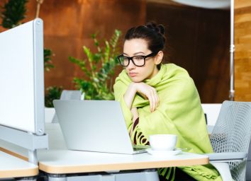woman coveted with coverlet using laptop and feeling cold on workplace