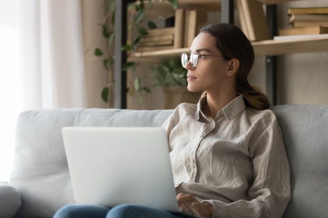 woman sit on couch hold laptop look in distance thinking distracted from online work