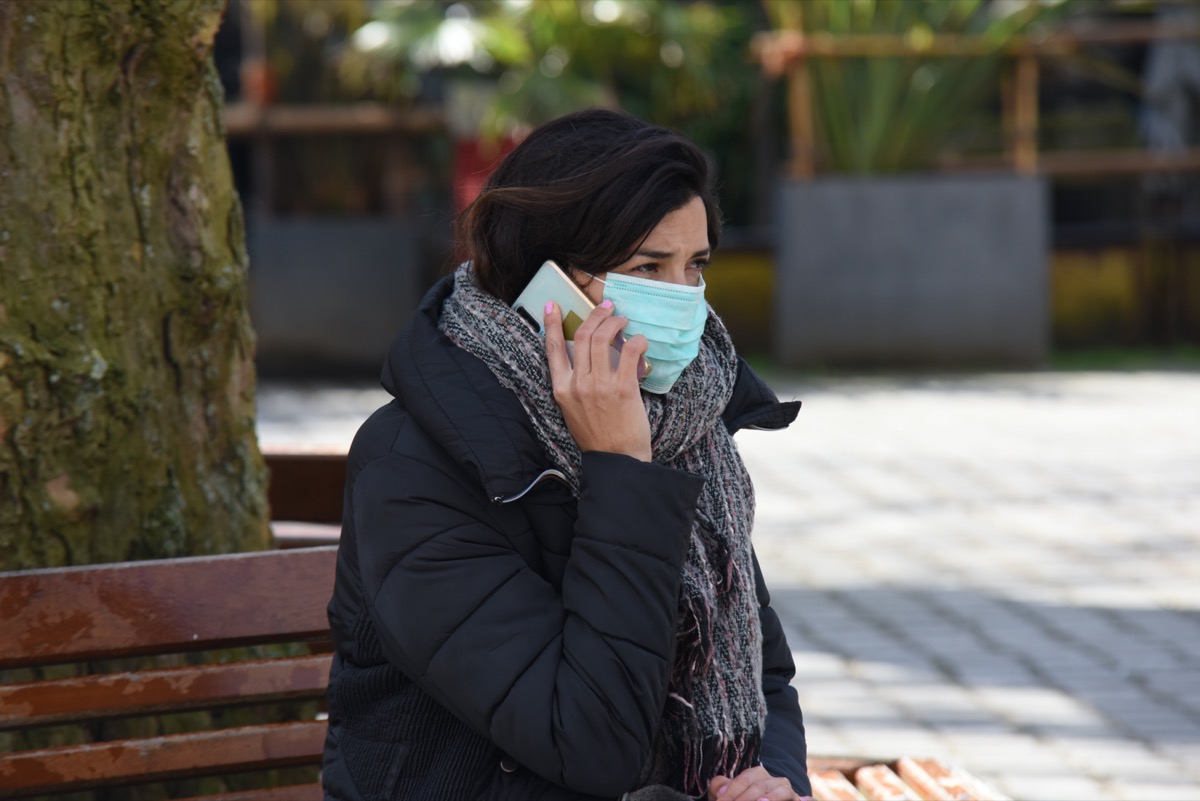 A brown hair girl sitting in a public park wearing protection mask calling friends and family for updates and news with her mobile phone