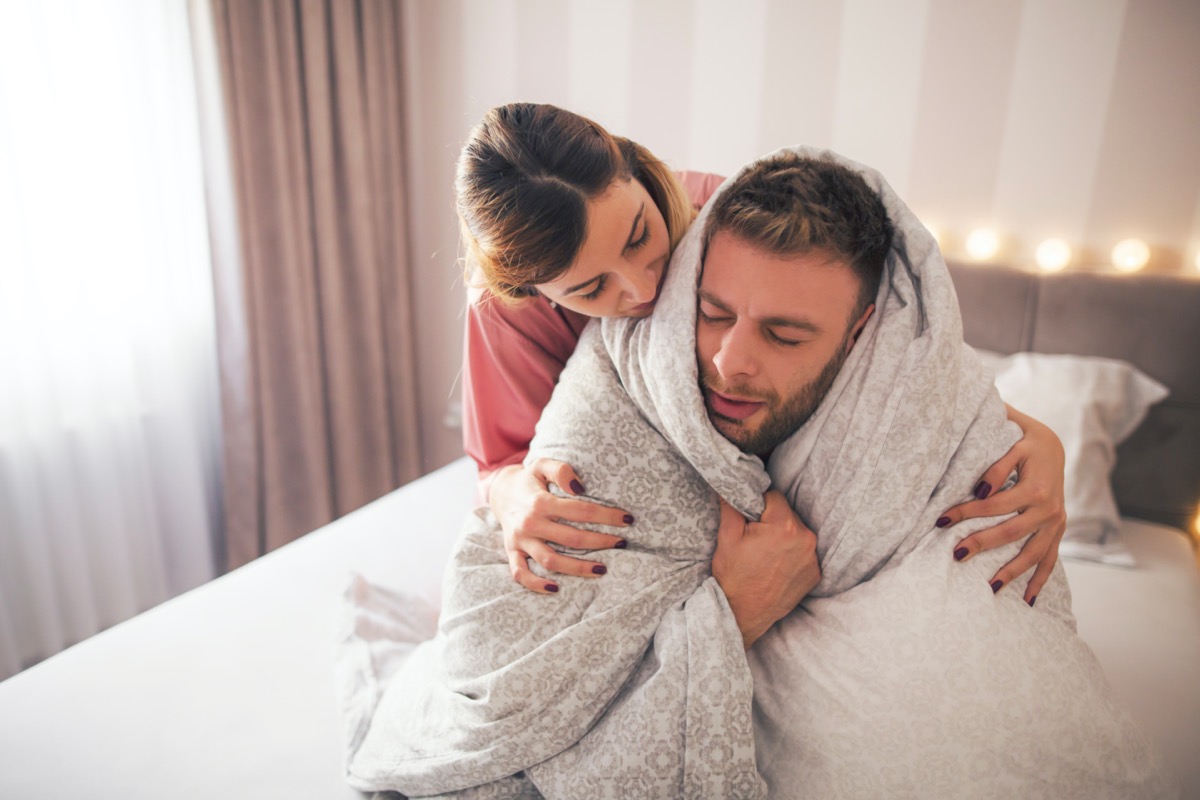 Man sitting on the bed wrapped in a blanket feeling sick while girl hugging him and trying to help him.