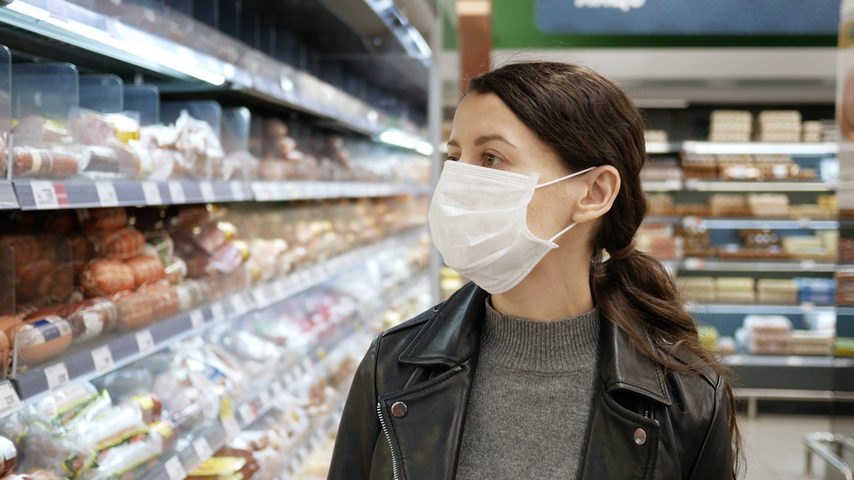 Young woman shopping in grocery store for food while wearing mask and preventing spread of coronavirus virus germs by wearing face mask.
