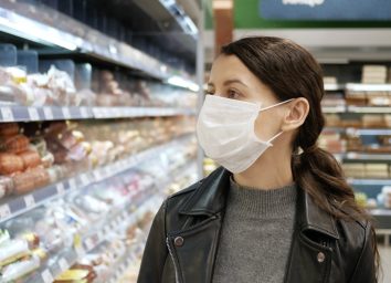 Young woman shopping in grocery store for food while wearing mask and preventing spread of coronavirus virus germs by wearing face mask.