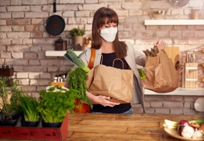 Woman wearing white medical face mask to prevent infection, arriving home, holding paper shopping bag. Protection against coronavirus