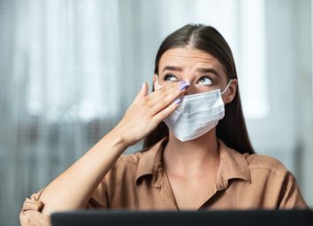 Don't Touch Your Face. Girl wearing surgical mask rubbing her eye with dirty hands, working on laptop