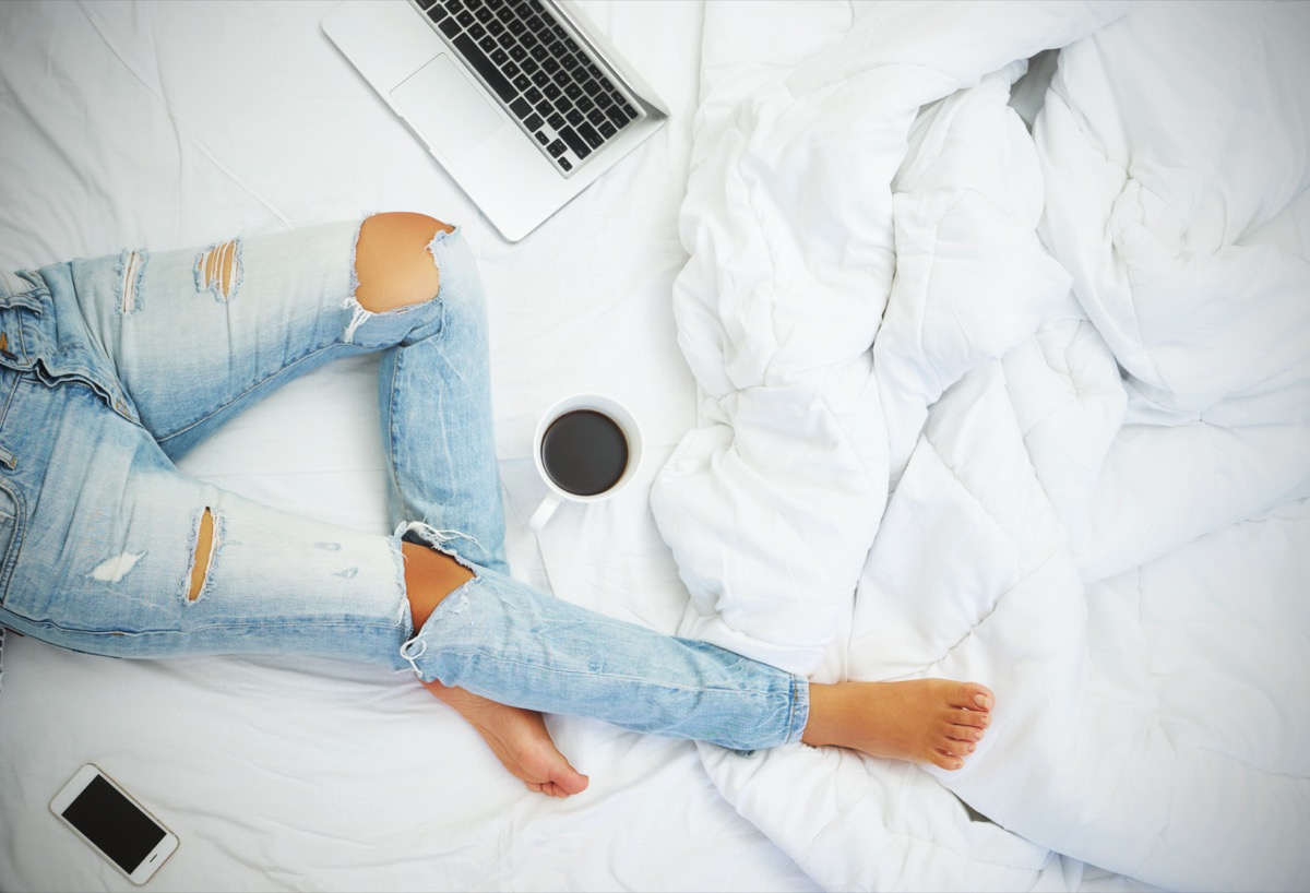 Young woman drinking coffee at home in her bed and checking her laptop, top view