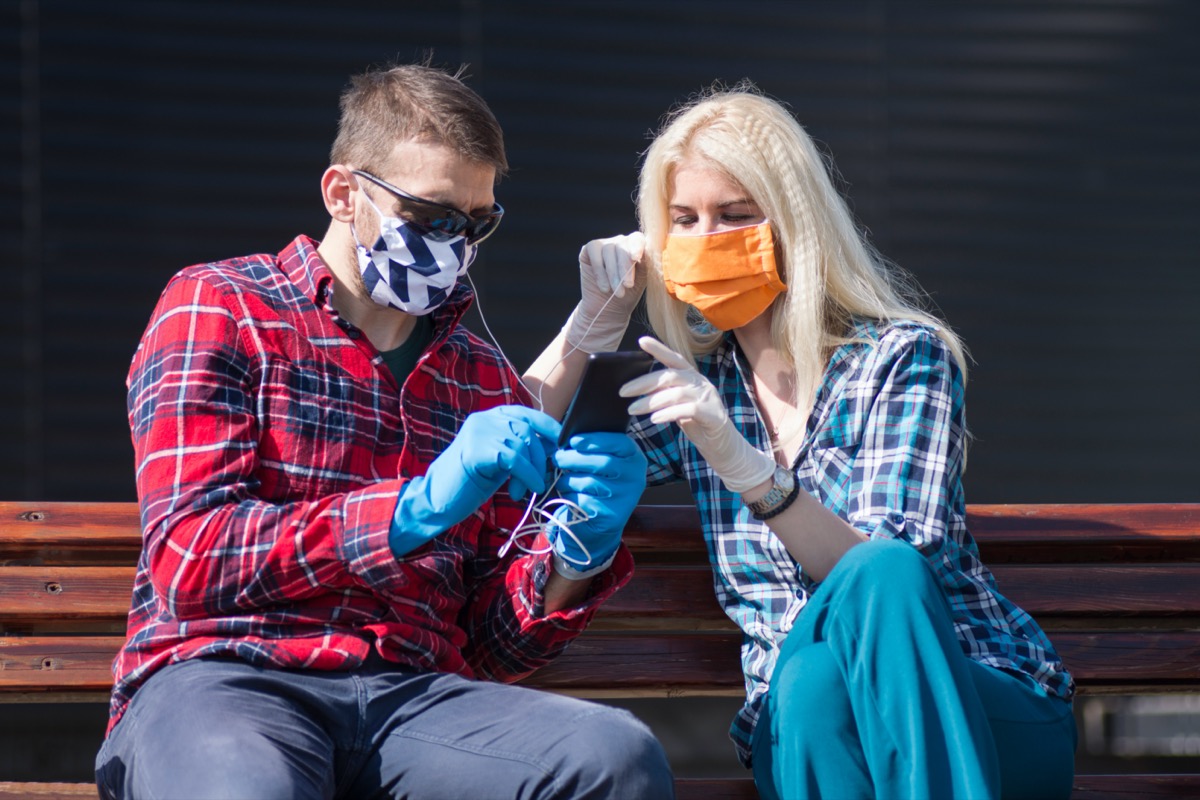 The couple with protective masks and gloves is listening music and using phone outdoors, modern lifestyle concept in coronavirus season.